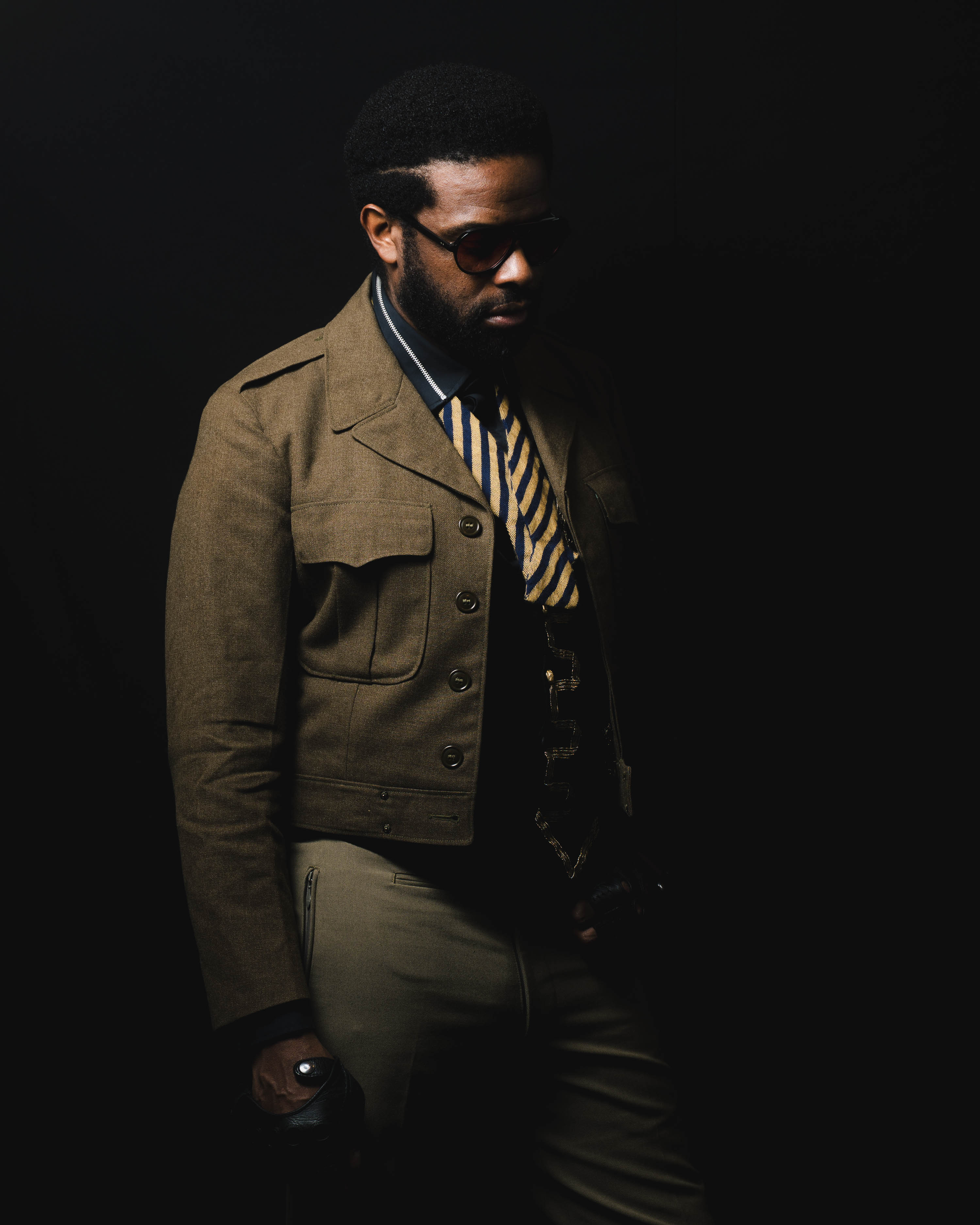 A photo portrait of Adrian Younge. He's standing in front of a black background while looking at the ground. He wears sunglasses, a green jacket and a tie with gold embroidered details.